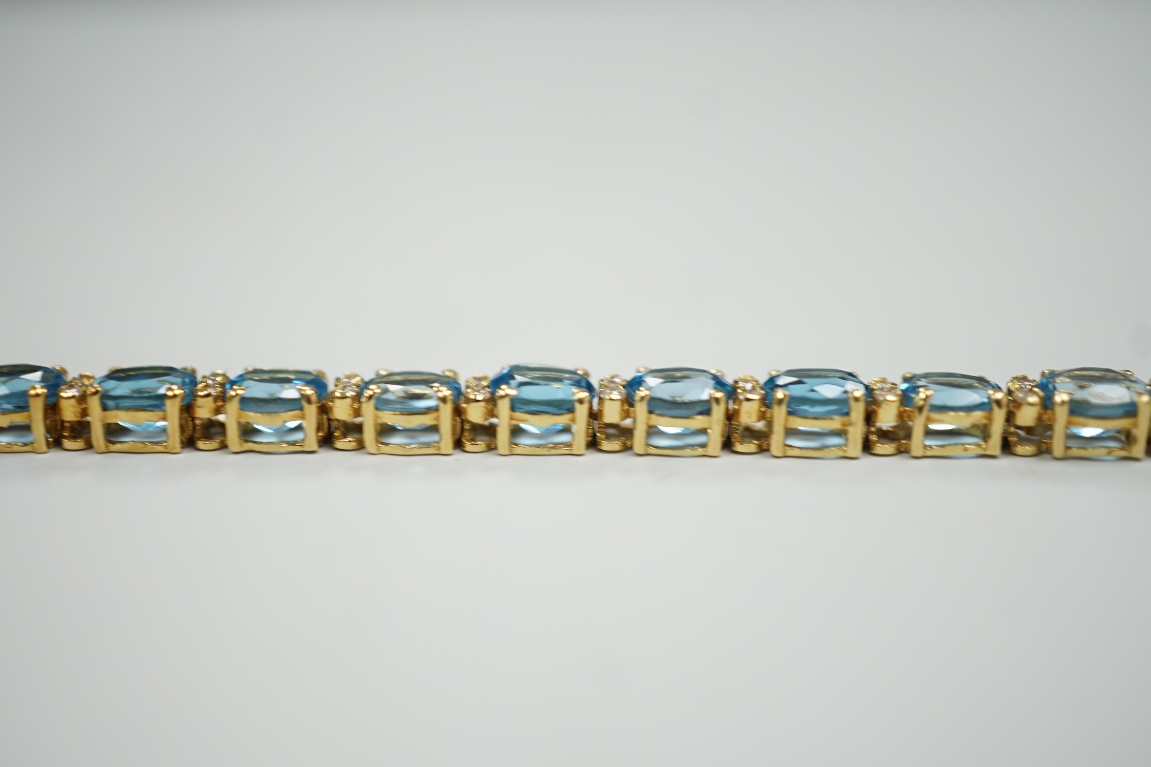 A modern 750 yellow metal and blue topaz set line bracelet, with diamond chip spacers, 17.75cm